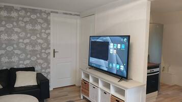 En stue med et fladskærms-tv på et stativ, en sofa, et sofabord og en dør med blomstret tapet.