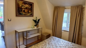 Ein Schlafzimmer mit einem Bett, einem Nachttisch mit Pflanze, einem Bild an der Wand und einem Fenster mit Vorhängen.
