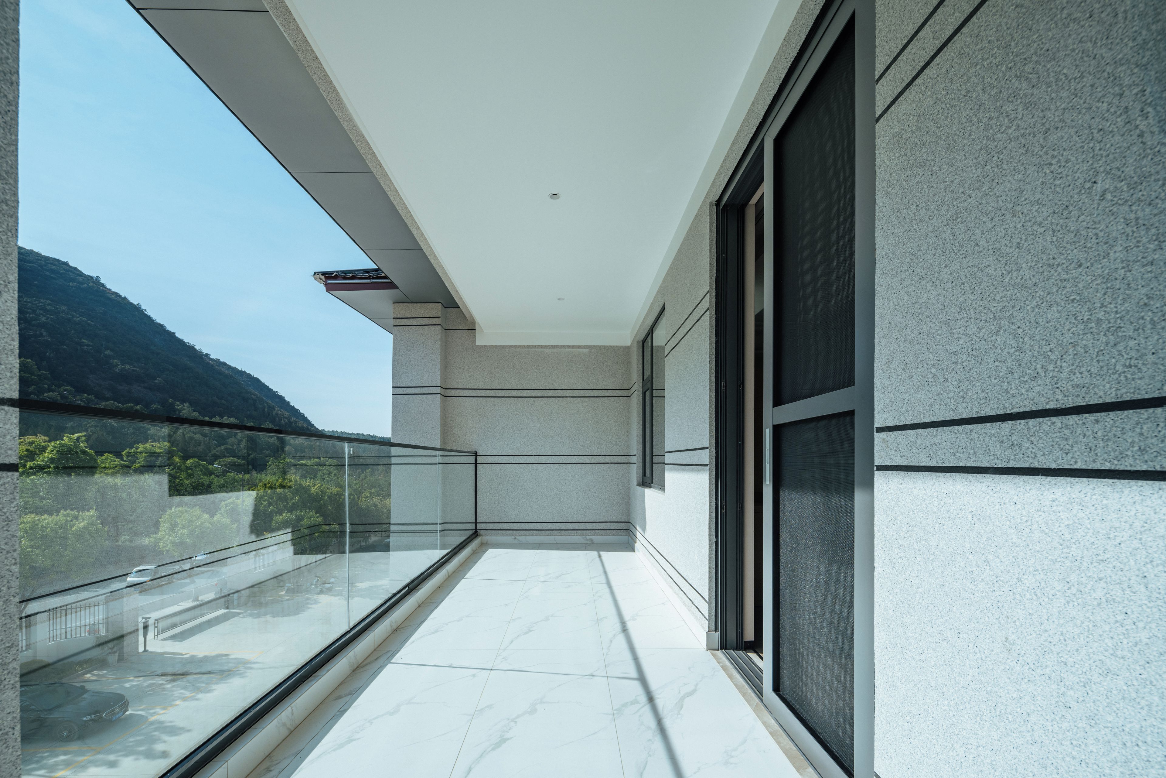 A modern balcony with a glass railing, a view of a mountain, and a building with a textured facade.