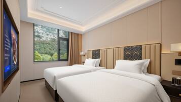 A hotel room with two beds, a large window with a view of greenery, and a flat-screen TV on the wall.
