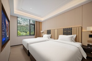 A hotel room with two beds, a large window with a view of greenery, and a flat-screen TV on the wall.
