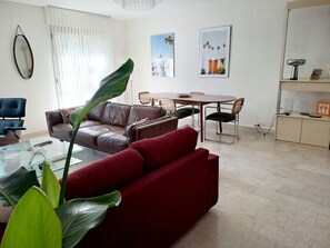 Ein Wohnzimmer mit braunem Ledersofa, einem roten Sofa, einem Glastisch und einem Essbereich mit Tisch und Stühlen.