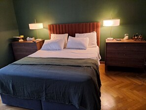 Een netjes opgemaakt bed met een donkerblauwe deken en witte kussens, aan weerszijden geflankeerd door nachtkastjes met lampen, in een kamer met groene wanden en houten vloeren.