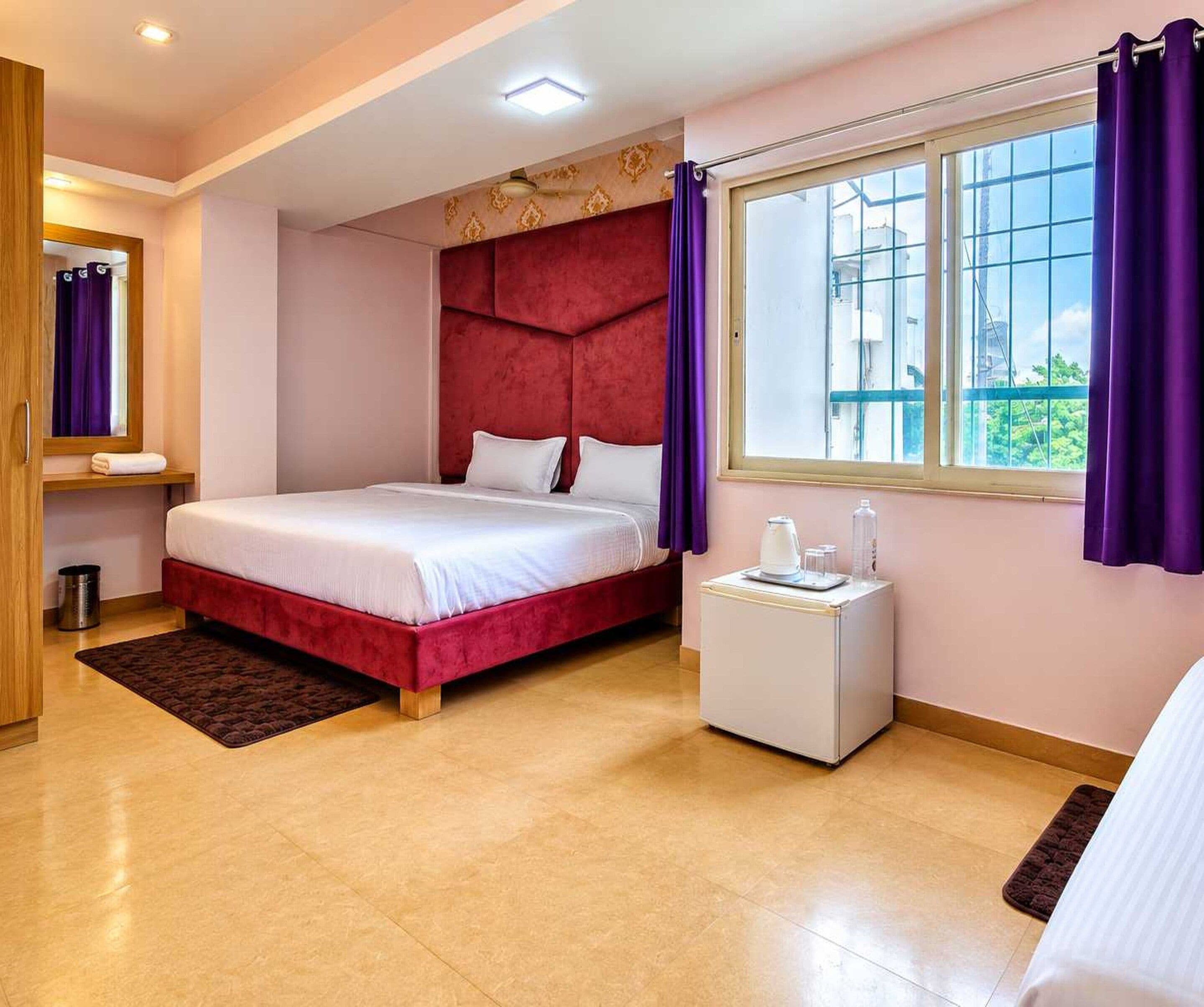 A hotel room with a bed, a small bedside table with a kettle, a window with curtains, and a mirror.