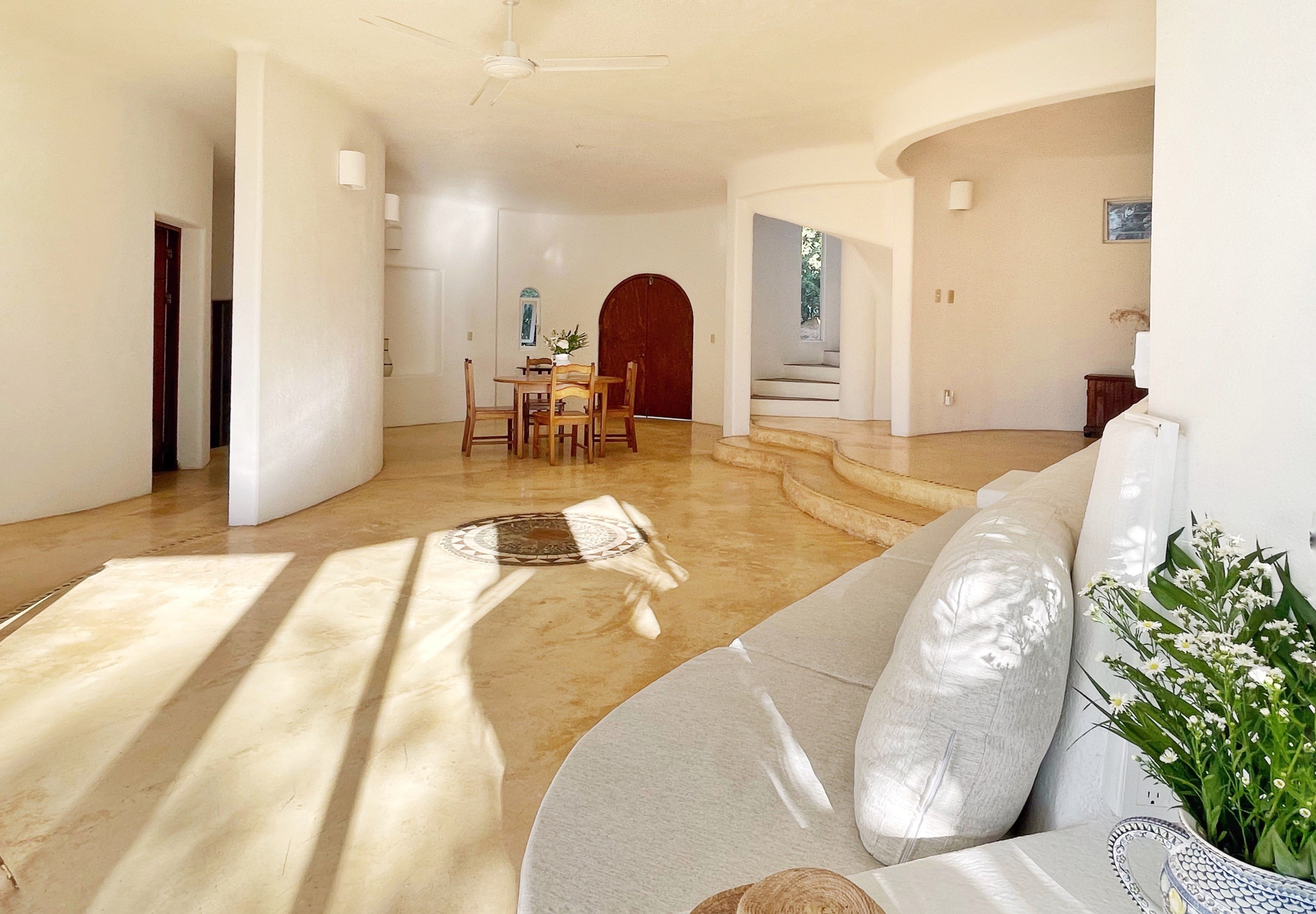 Uma sala de estar ampla, com um sofá grande, uma mesa de jantar com cadeiras e uma escada que dá acesso a um andar superior.