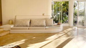 Sala de estar moderna com sofá curvo, mesinha lateral e vista para um jardim exuberante através de grandes janelas.