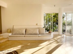 Ein modernes Wohnzimmer mit einer gebogenen Couch, einem kleinen Beistelltisch und Blick auf einen üppigen Garten durch große Fenster.