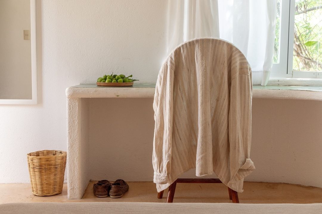 A room with a chair covered in a white cloth, a small table with a bowl of green apples, a wicker basket, and a pair of shoes on the floor.