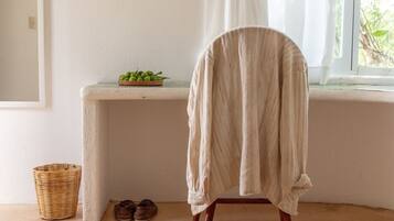 A room with a chair covered in a white cloth, a small table with a bowl of green apples, a wicker basket, and a pair of shoes on the floor.