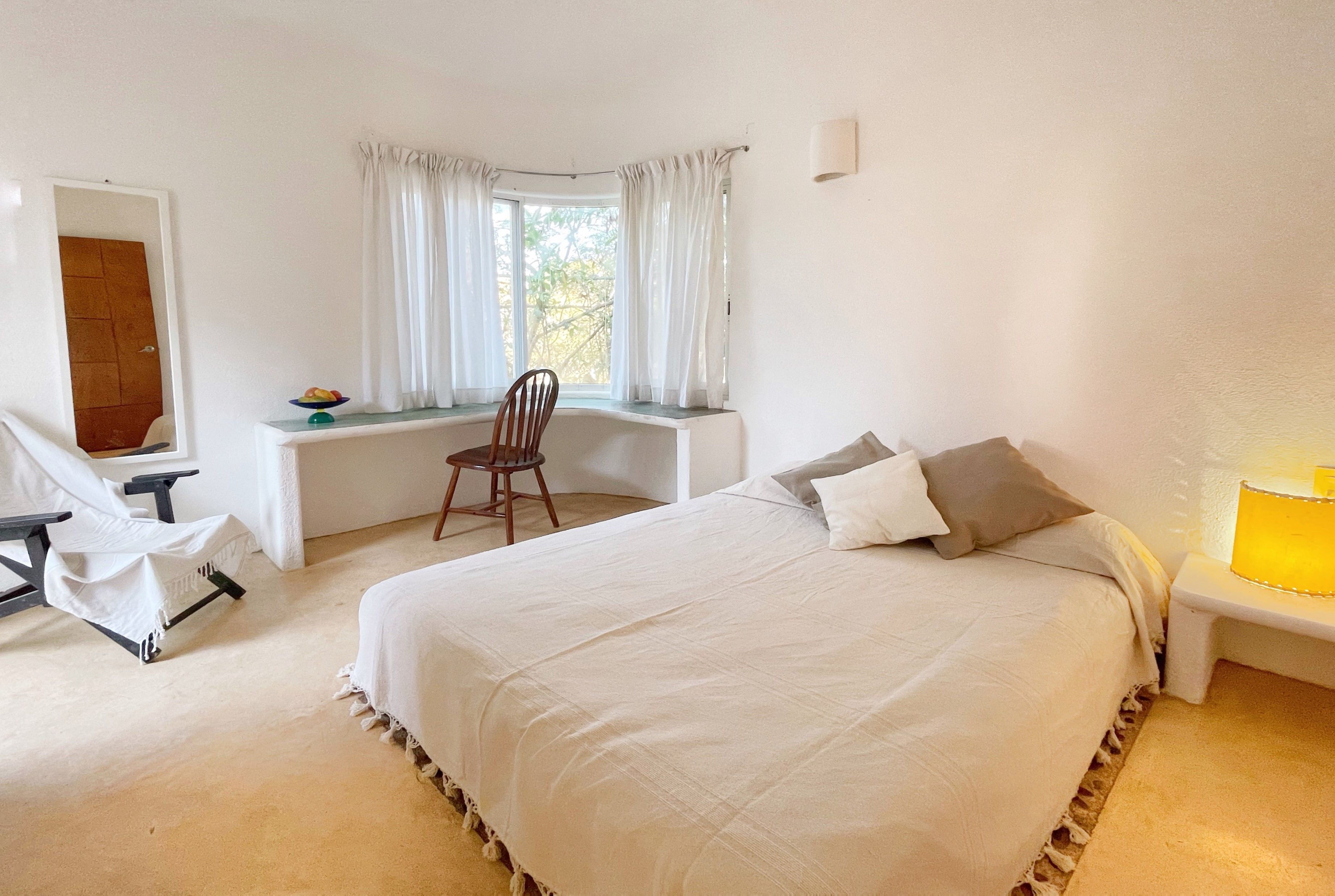 A bedroom with a bed, a desk, a chair, a window with curtains, and a mirror.