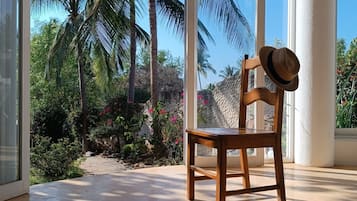 Uma cadeira de madeira com um chapéu, colocada perto de uma grande janela com vista para palmeiras e um jardim.
