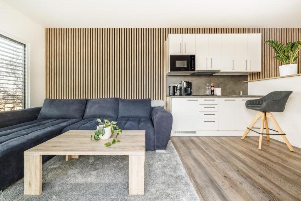Un espace de vie moderne avec un canapé foncé, une table basse en bois, un coin cuisine doté de meubles blancs et une plante.