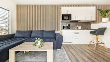 Un espace de vie moderne avec un canapé foncé, une table basse en bois, un coin cuisine doté de meubles blancs et une plante.