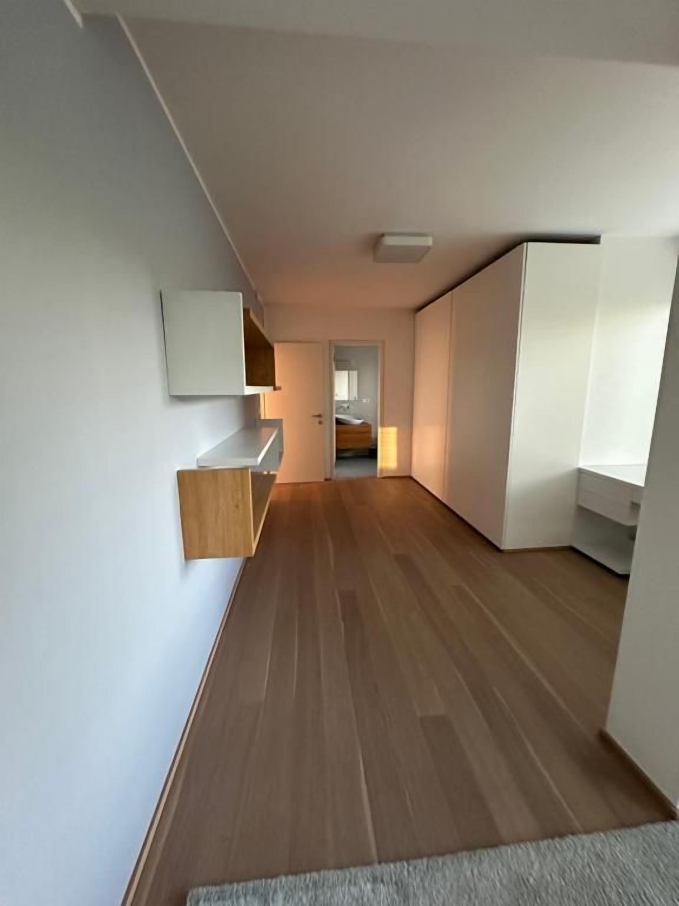 Un couloir étroit avec un plancher en bois, des murs blancs et une porte donnant sur une autre pièce.
