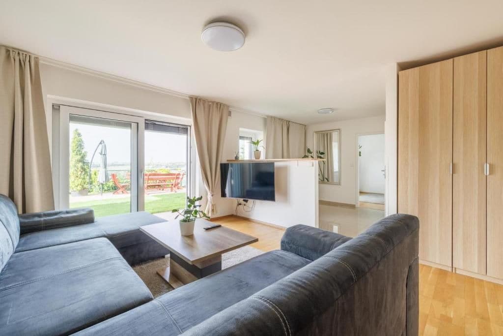 A modern living room with a sofa set, a flat-screen TV, and a sliding glass door leading to a balcony.