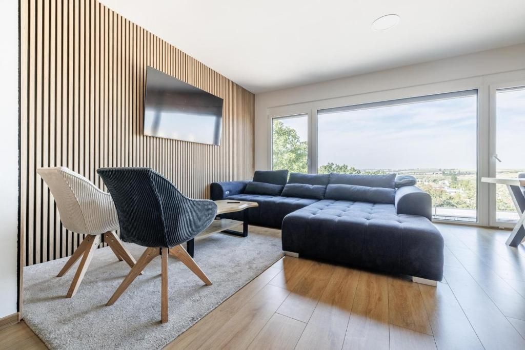 A modern living room with a sectional sofa, a black chair, a flat-screen TV mounted on a wooden wall, and large windows offering a view.