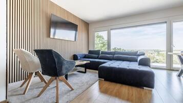A modern living room with a sectional sofa, a black chair, a flat-screen TV mounted on a wooden wall, and large windows offering a view.