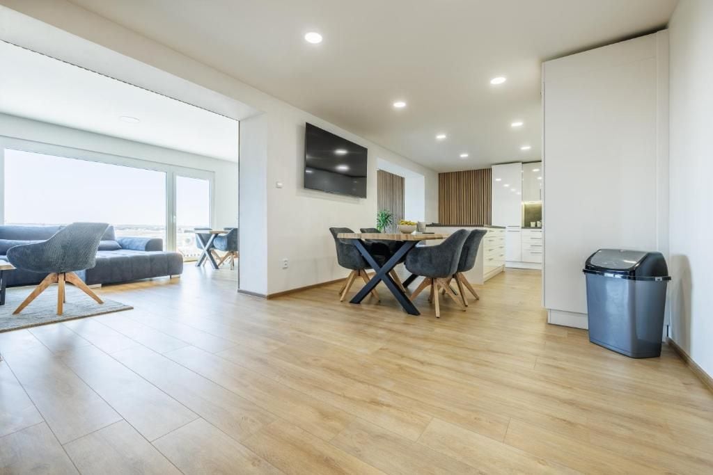 A modern living room with a sofa, dining table, and chairs. There is a TV mounted on the wall and a trash bin against the wall.