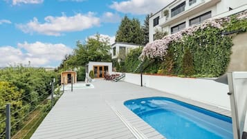 A modern outdoor swimming pool area with a wooden deck, a small building, and a white building with a garden.