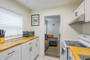 A compact kitchen with white cabinets, a microwave, and a stove. There's a window with blinds, a framed picture on the wall, and a small rug leading to a living area. - 2 Mi to Dtwn Savannah! WFH-Friendly Retreat (Savannah)