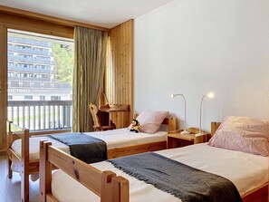 A room with two wooden beds, a window with curtains, a view of a building labeled 'VICTORIA RESIDENCE,' and a small table with a lamp. - Les Dryades 8 by Interhome (Villars)