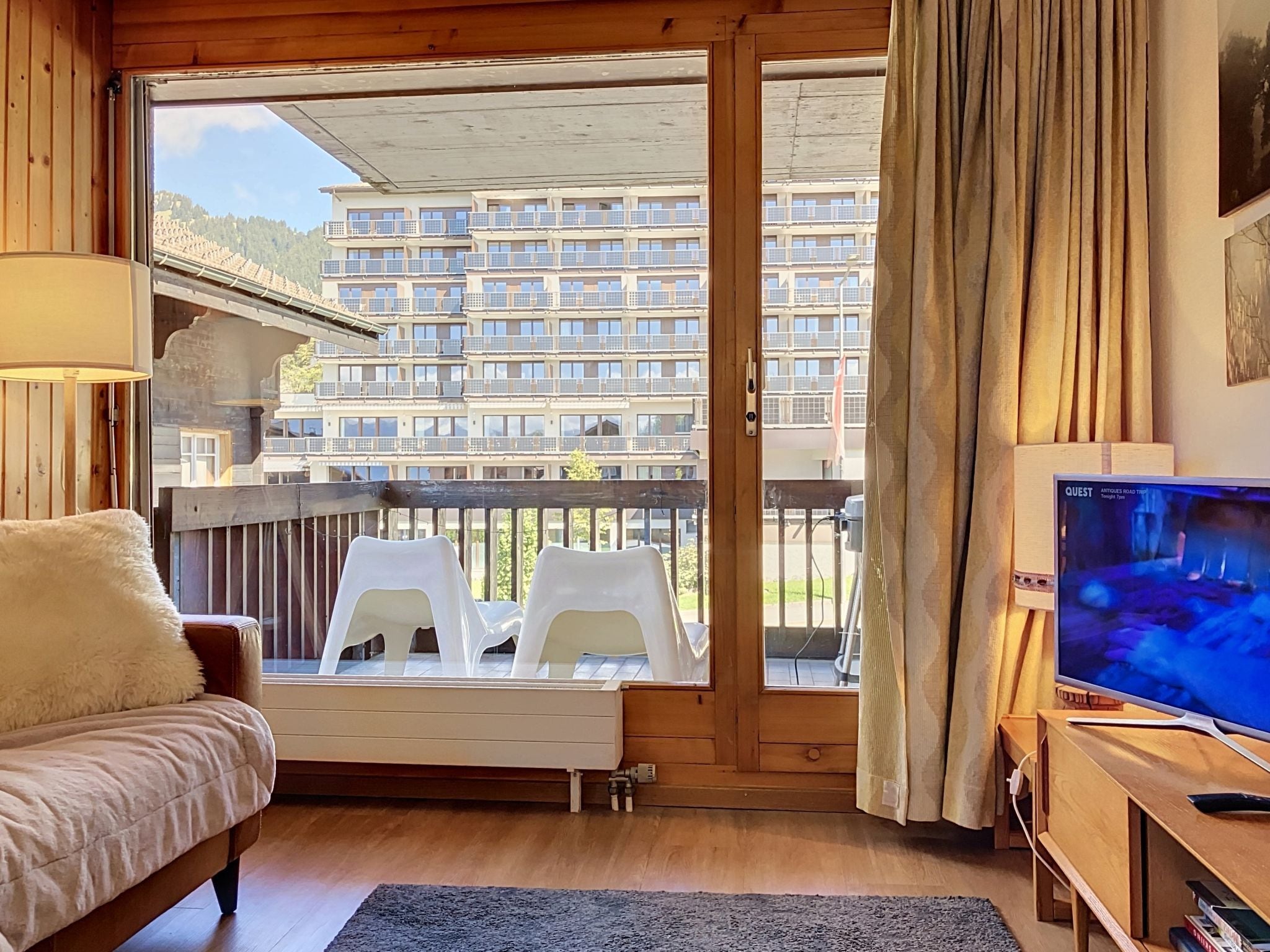 A room with a sofa, a TV, a balcony with plastic chairs, and a view of a multi-story building.