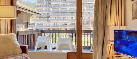 Une pièce avec un canapé, un téléviseur, un balcon avec des chaises en plastique et une vue sur un immeuble à plusieurs étages.