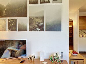 Un salon avec une télévision, des tableaux accrochés au mur, une petite table avec une télécommande, un haut-parleur blanc et un sapin de Noël.