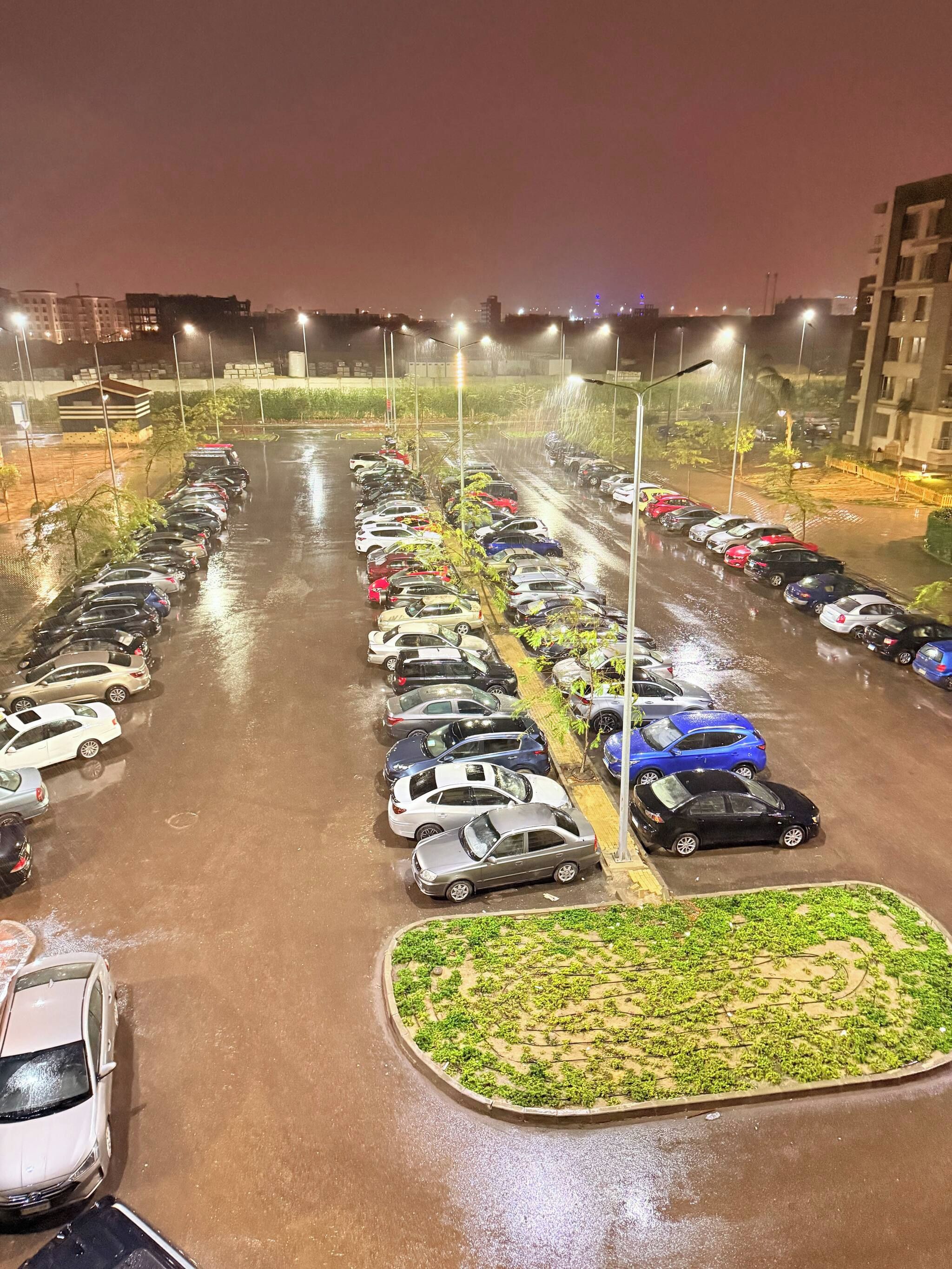A parking lot with multiple rows of parked cars, illuminated by overhead lights.