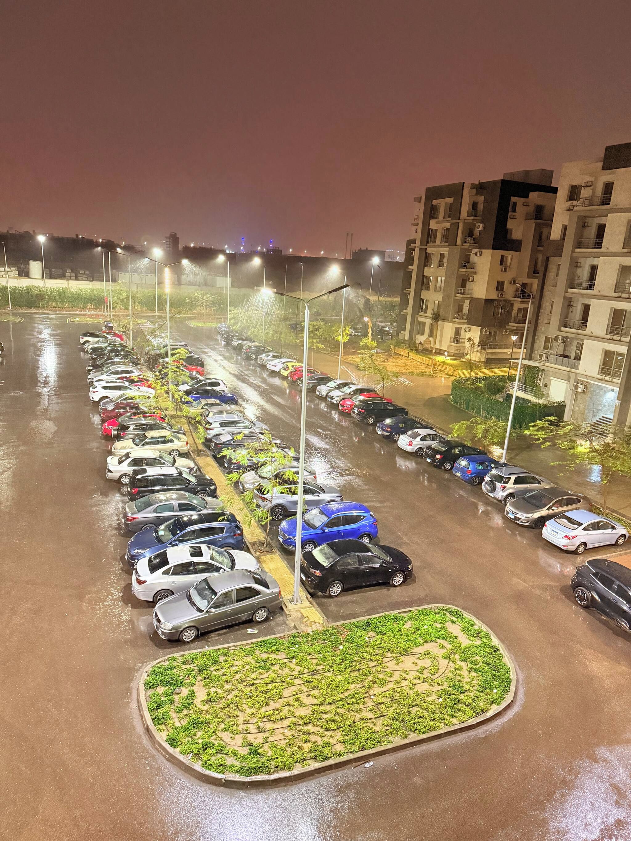 Un estacionamiento nocturno con coches aparcados en filas, rodeado de edificios y farolas.
