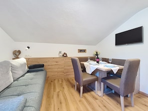 A cozy dining area with a wooden table set for two, a grey sofa, and a television mounted on the wall. - Apartment Enzian by Interhome (Mayrhofen)