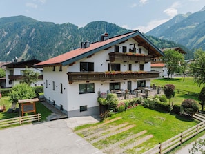 A two-story building with a red-tiled roof, surrounded by lush greenery and mountains. - Apartment Enzian by Interhome (Mayrhofen)