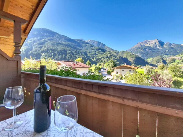 En balkon med et bord, en flaske og to glas, med udsigt over et bjergrigt landskab med huse.