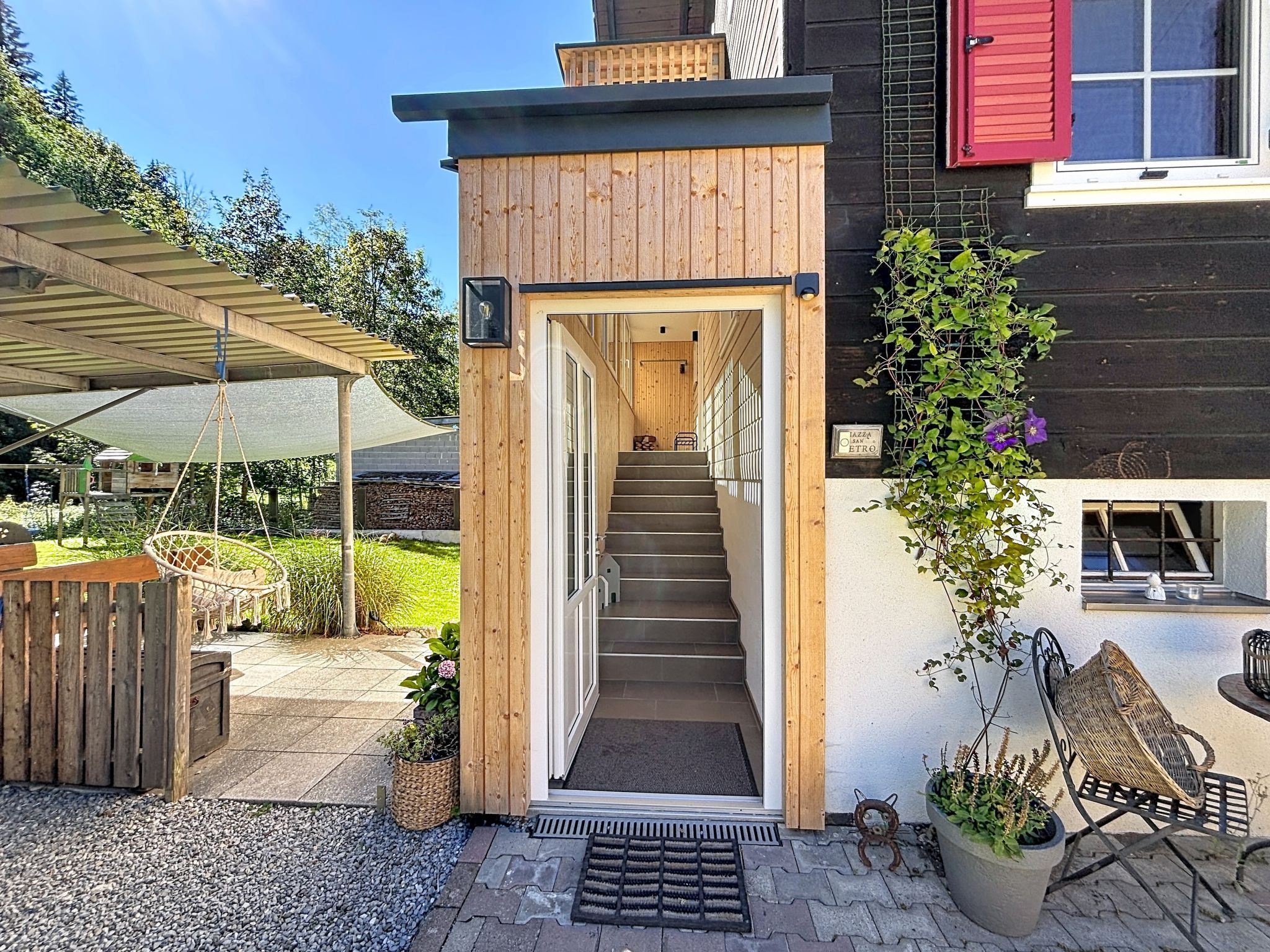 Fachada de madera con una escalera que lleva a una puerta, un área de estar al aire libre cubierta y un jardín con plantas y un banco.