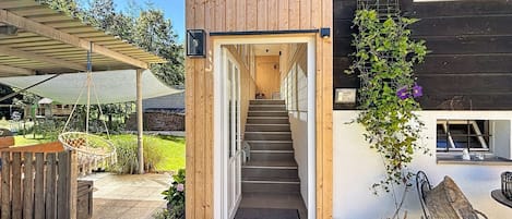 Fachada de madera con una escalera que lleva a una puerta, un área de estar al aire libre cubierta y un jardín con plantas y un banco.