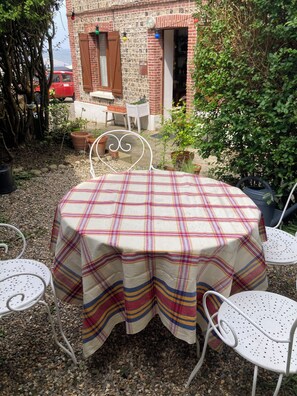A round table with a plaid tablecloth, surrounded by white metal chairs, set outdoors with a brick building and greenery in the background.