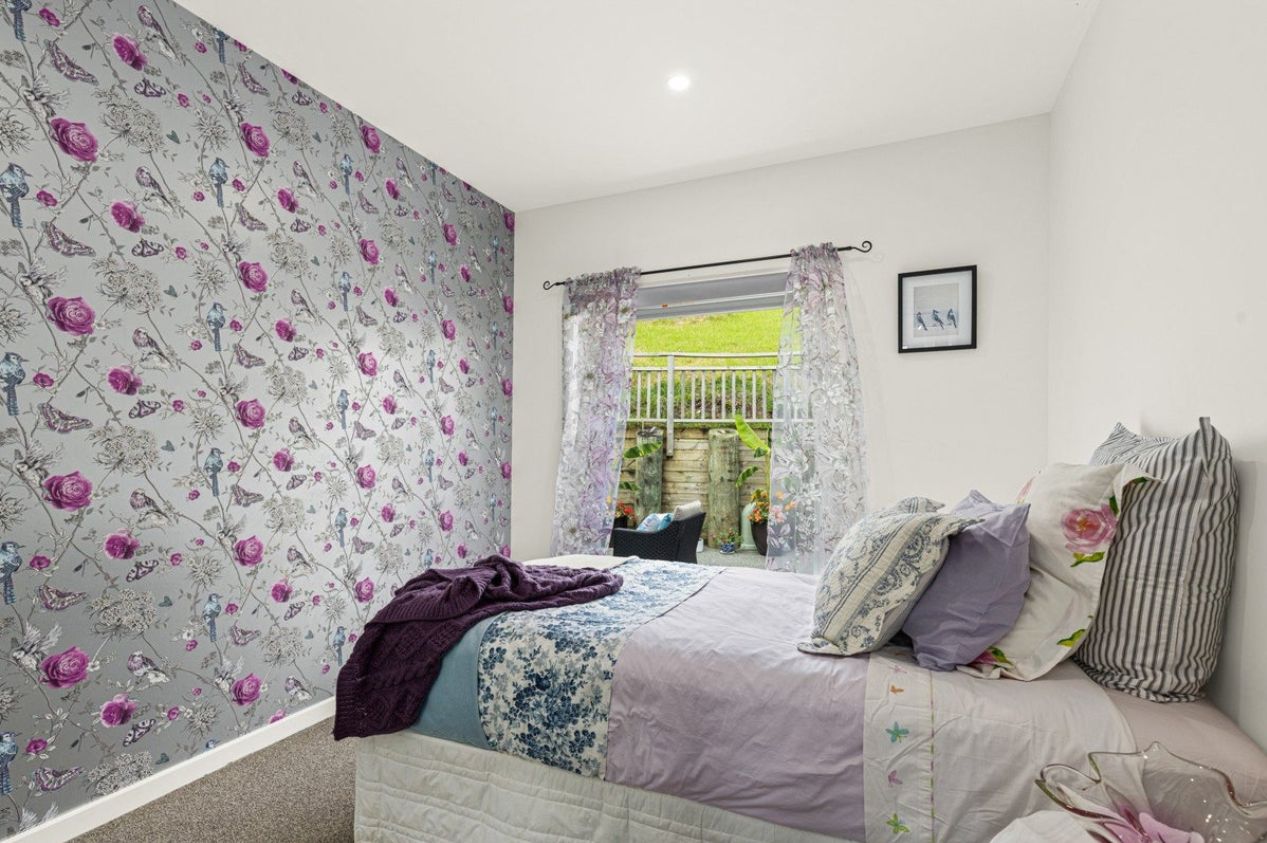 Ein Schlafzimmer mit blumigem Wandpapier, einem Bett mit vielen Kissen, einem Fenster mit Vorhängen und einem gerahmten Bild an der Wand.
