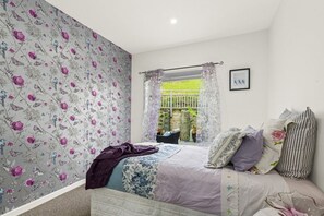Un dormitorio con papel tapiz floral, una cama con muchos cojines, una ventana con cortinas y un cuadro enmarcado en la pared.