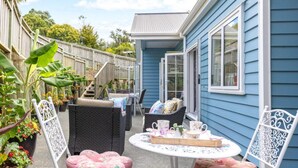 Een terras met rieten meubels, een tafel met een dienblad en een blauw huis op de achtergrond.