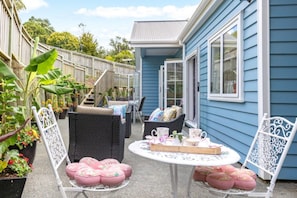 A patio with wicker furniture, a table with a tray of items, and a blue house in the background. - Villa Vie: Spacious family escape in Warkworth near Matakana (Warkworth)