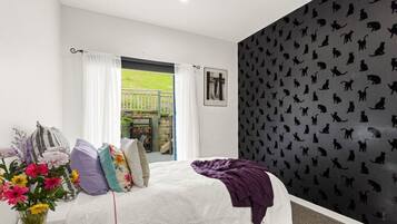 A bedroom with a bed, a window with curtains, a wall with a cat pattern, and a view of a garden.
