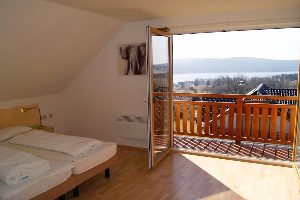 A bedroom with a bed, a nightstand, a window with a view of a body of water and mountains, and a balcony with a wooden railing.