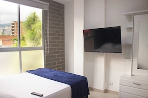 Una habitación de hotel con una cama, un televisor montado en la pared y una ventana con vistas a edificios y árboles.