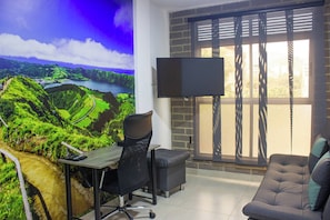 Una habitación con escritorio, silla de oficina y sofá. Hay un televisor montado en la pared y una ventana con persianas.