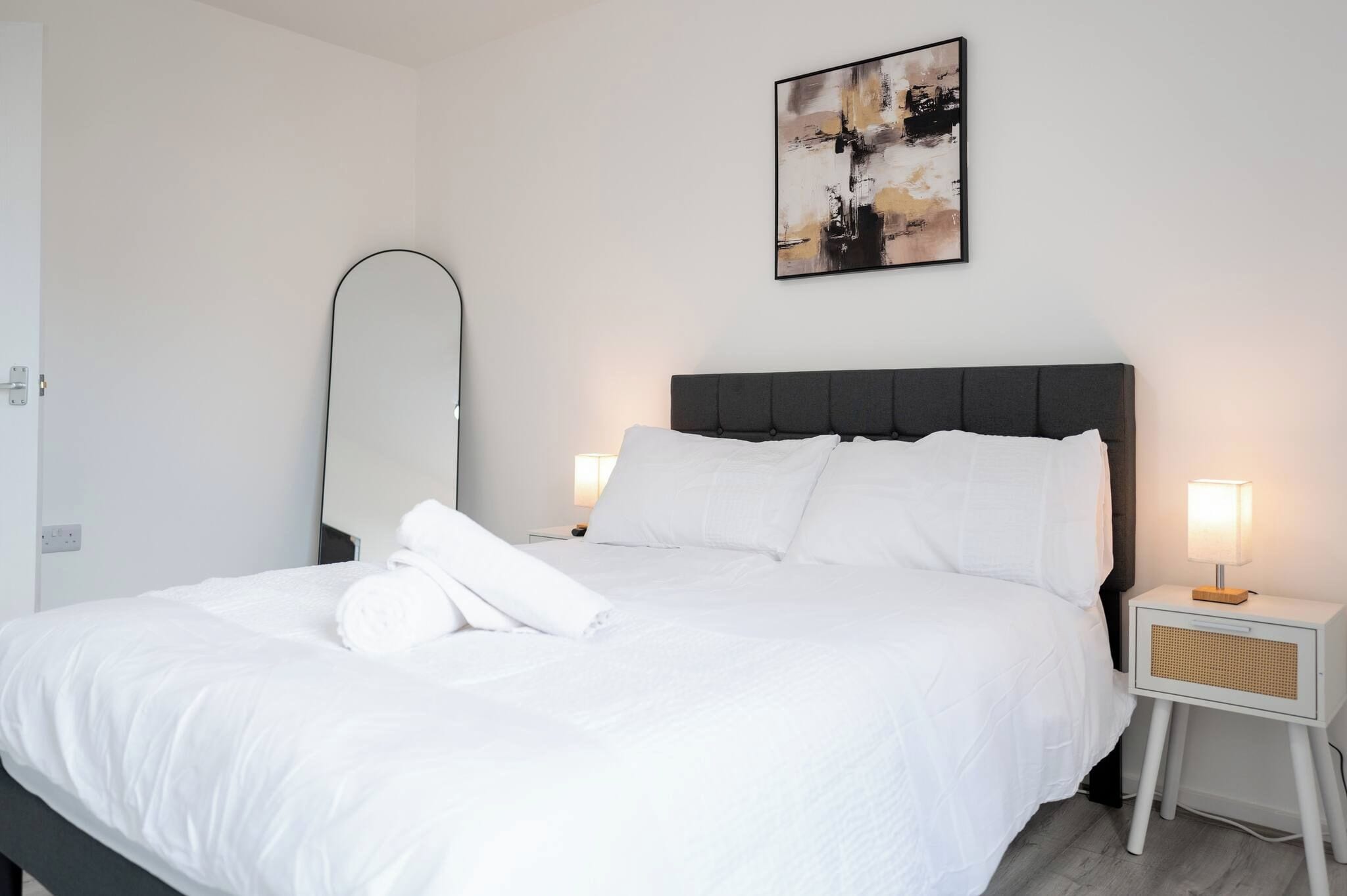 A hotel room with a bed, a bedside table with a lamp, a wall-mounted mirror, and a painting.