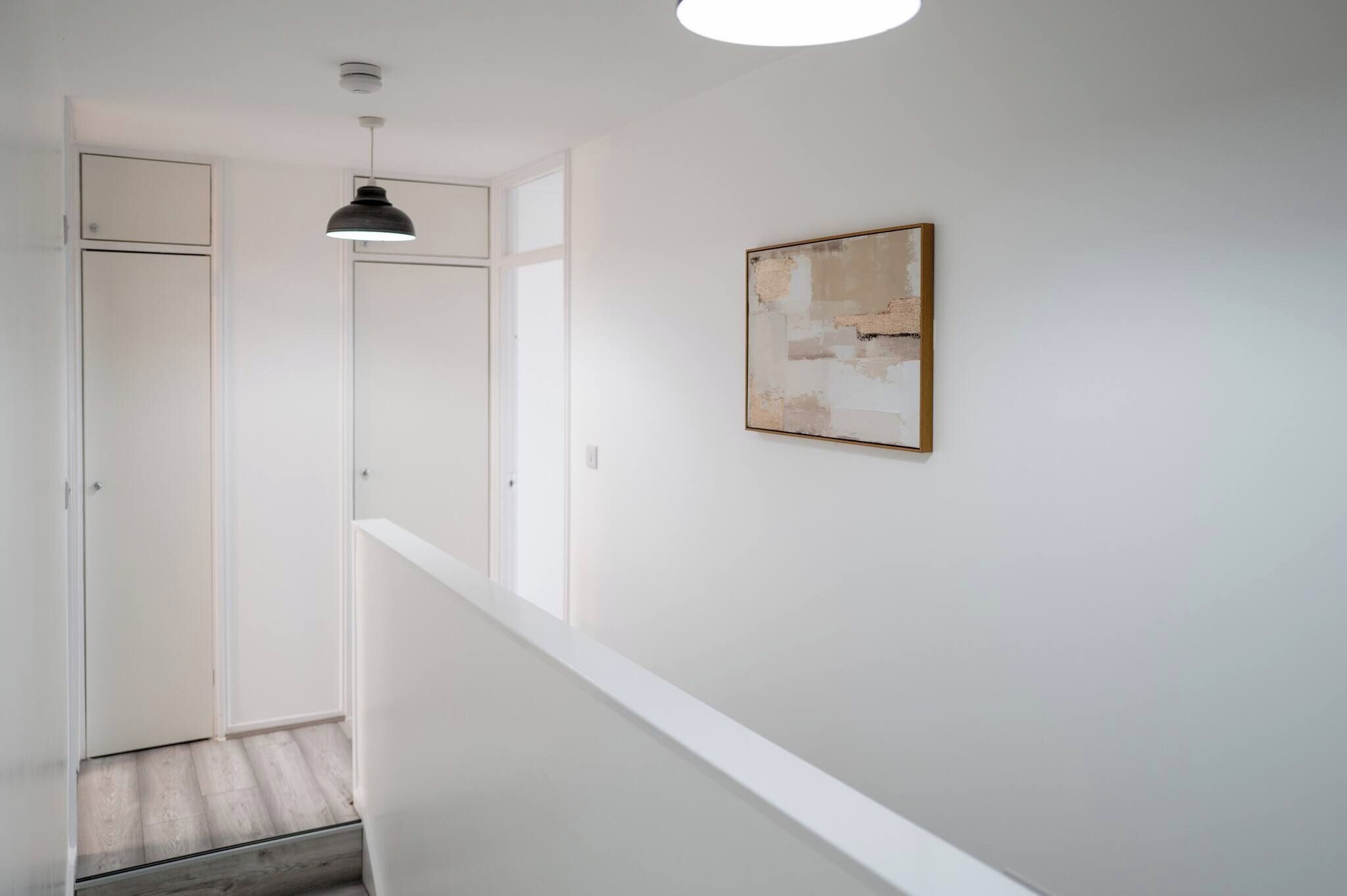 A minimalist hallway with a white railing, a pendant light, and a framed abstract painting on the wall.
