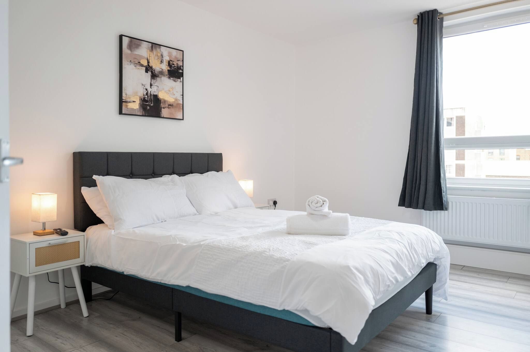 A neatly made bed with white bedding, a dark headboard, and a bedside table with a lamp.