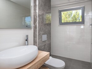 Ein modernes Badezimmer mit einem großen, ovalen Waschbecken, einer Toilette und einem Fenster mit Blick auf Bäume.