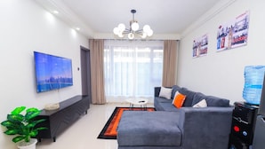 A modern living room with a grey sofa, a flat-screen TV, a potted plant, and a water dispenser. - Spacious 1-bedroom apartment in Westlands, Nairobi, Kenya (Nairobi)