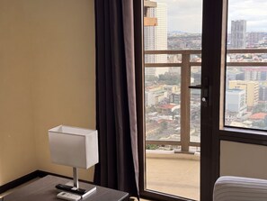 A hotel room with a view of a cityscape through a large window, a bedside table with a lamp, and a remote control. - Premier Double Room with Balcony and Manila Bay View, Pool View or MOA View (Pasay City)